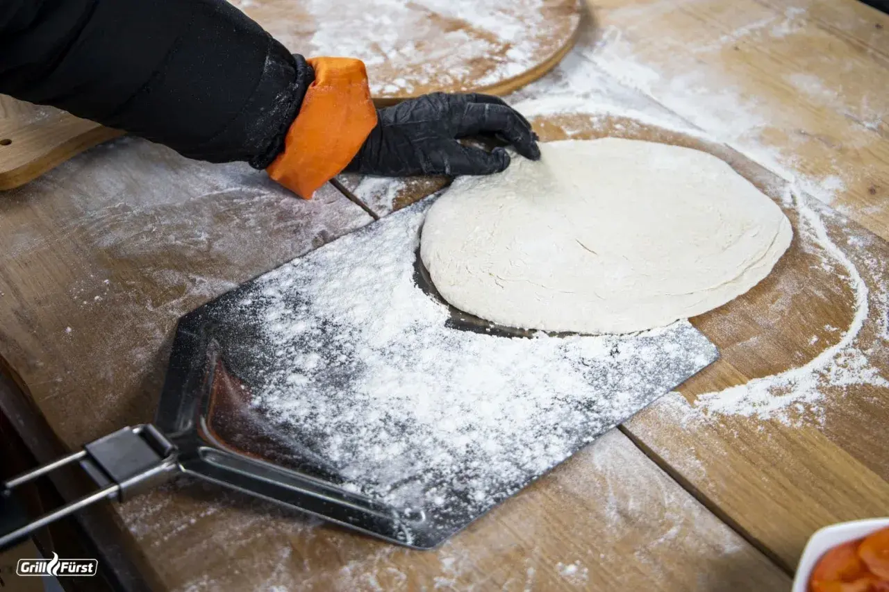 Hand mit Handschuh legt Teig auf bemehltes Pizzaschaufel.