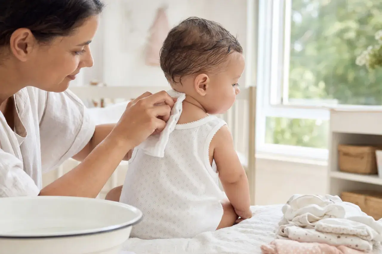 Madre limpia la nuca de su bebé para prevenir la sudamina. El bebé, sentado, mira hacia la ventana.