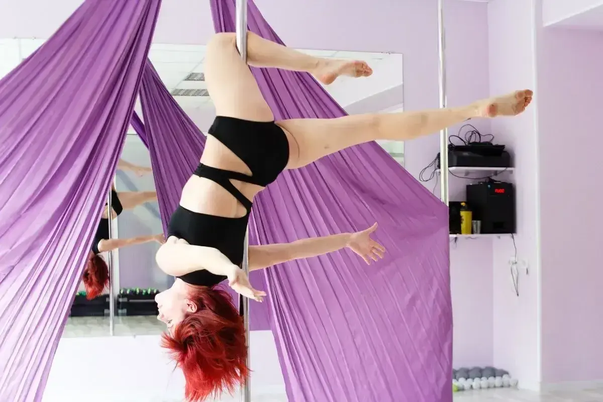 Kobieta z rudymi włosami wykonuje figurę pole dance invert, zwisając z rurki.