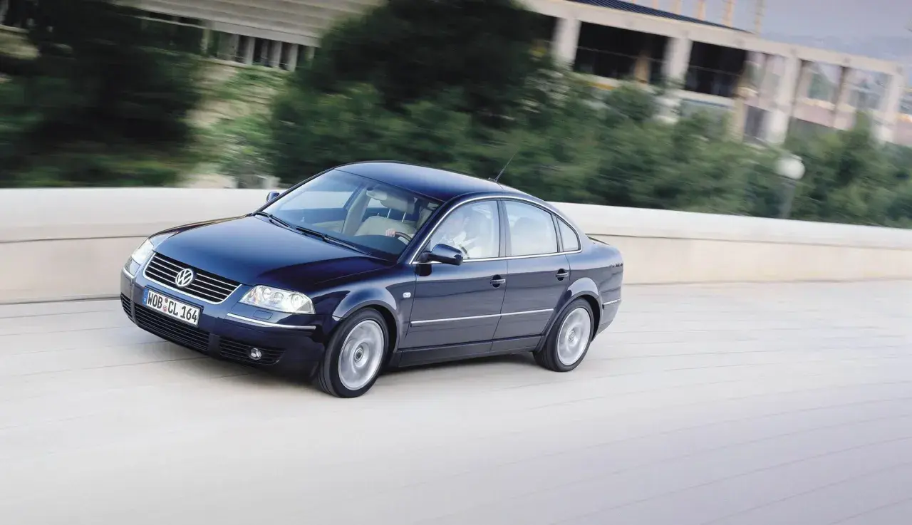 Ciemnoniebieski Volkswagen Passat B5 jedzie po moście, za nim rozmyte drzewa i budynek.