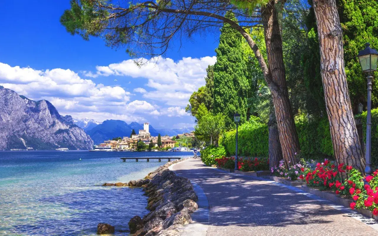 Promenade ensoleillée au bord du lac de Garde, avec des montagnes majestueuses en arrière-plan et un village pittoresque.