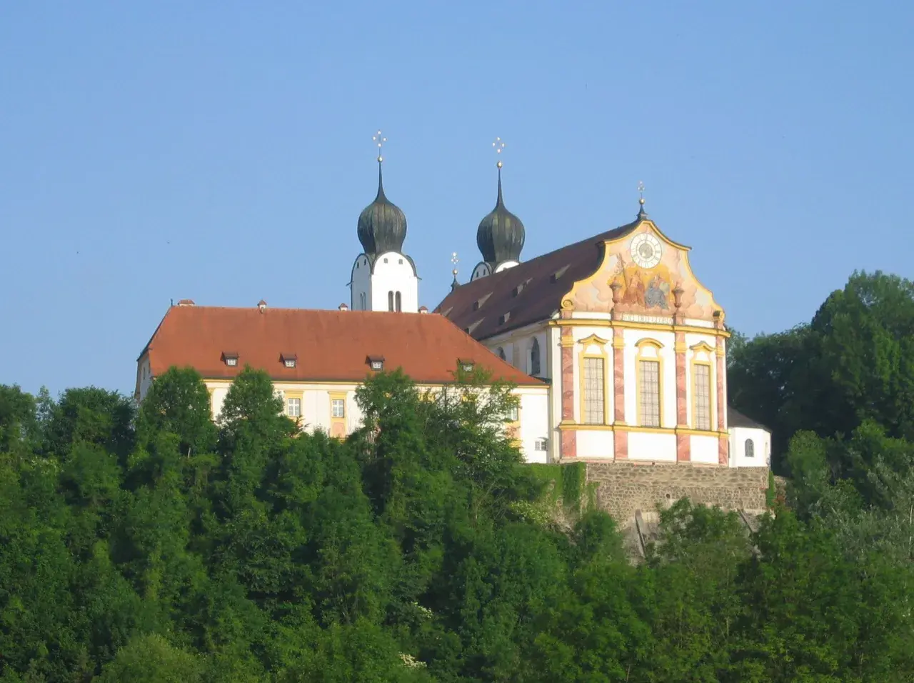 Prachtvolles Kloster Baumburg thront auf einem Hügel, umgeben von üppigem Grün. Zwei markante Zwiebeltürme ragen in den blauen Himmel.