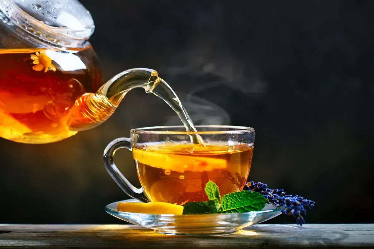 Un thé ambré, aux reflets de la noire, est versé d'une théière en verre dans une tasse ornée de citron et de menthe.