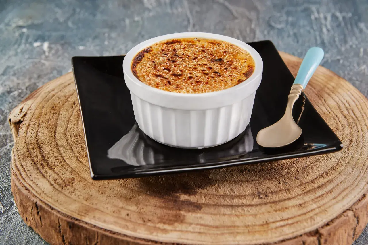 Crème brûlée avec une croûte caramélisée dorée, servie dans un ramequin blanc sur une assiette noire, avec une cuillère à la touche champignon.