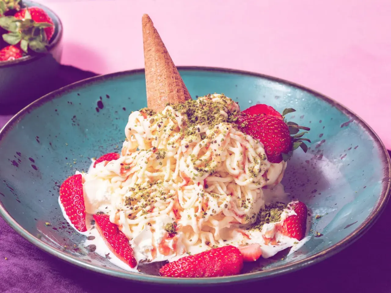 Spaghetti-Eis mit Erdbeeren und Pistazien in einer blauen Schale, garniert mit einer Eistüte.