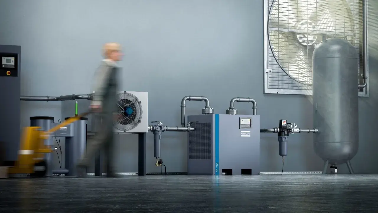 Industrielle Anlage mit Kompressor, Trockner und Luftbehälter. Ein Mitarbeiter mit Hubwagen im Vordergrund. Regelmäßiges kompressor wasser ablassen ist wichtig für die Effizienz.