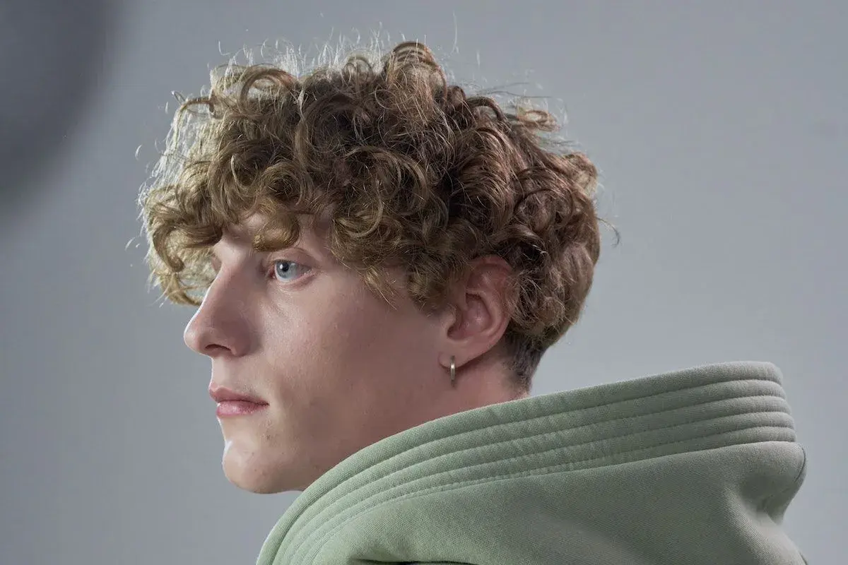 Primo piano di un giovane con capelli mossi uomo, occhi azzurri e un orecchino a cerchio. Indossa una felpa verde chiaro.