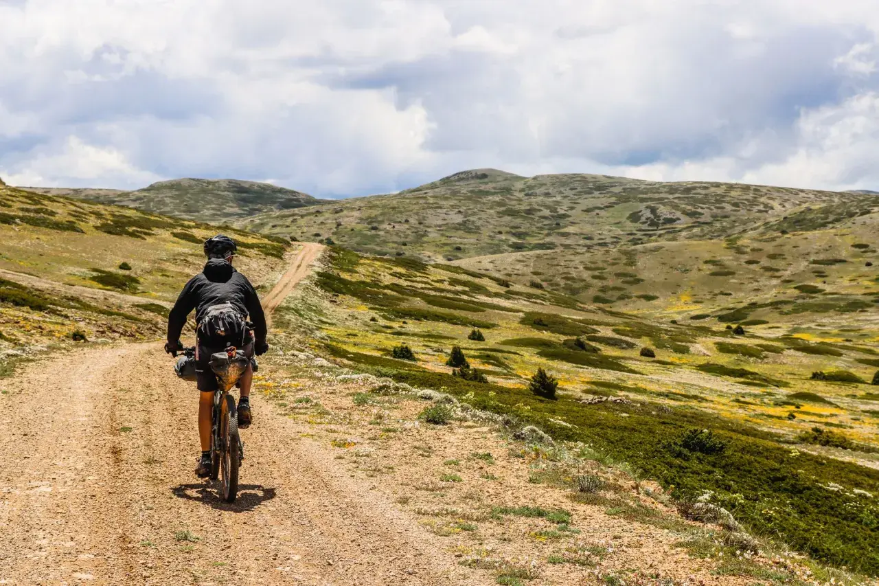 Ciclista pedaleando por un camino de tierra en **montañas vacías** bajo un cielo nublado.