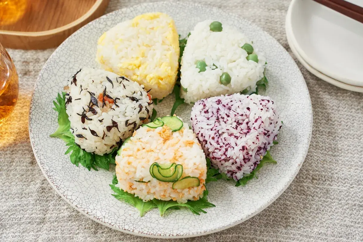 A colorful assortment of musubi vs onigiri, each a unique rice ball shape with various toppings like peas, seaweed, and cucumber.
