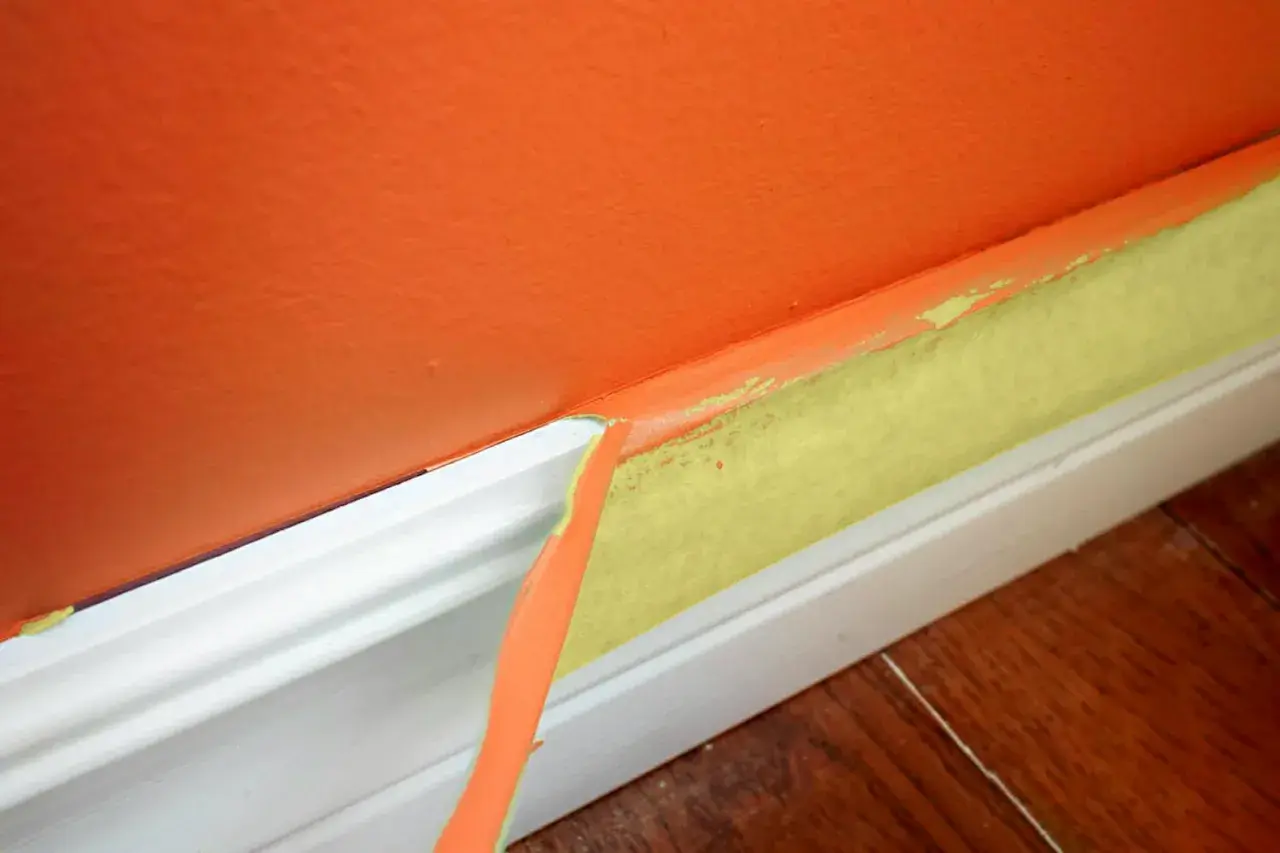 Ta bort maskeringstejp efter målning ger en ren kant. Orange vägg möter vit golvlist med gul tejp.