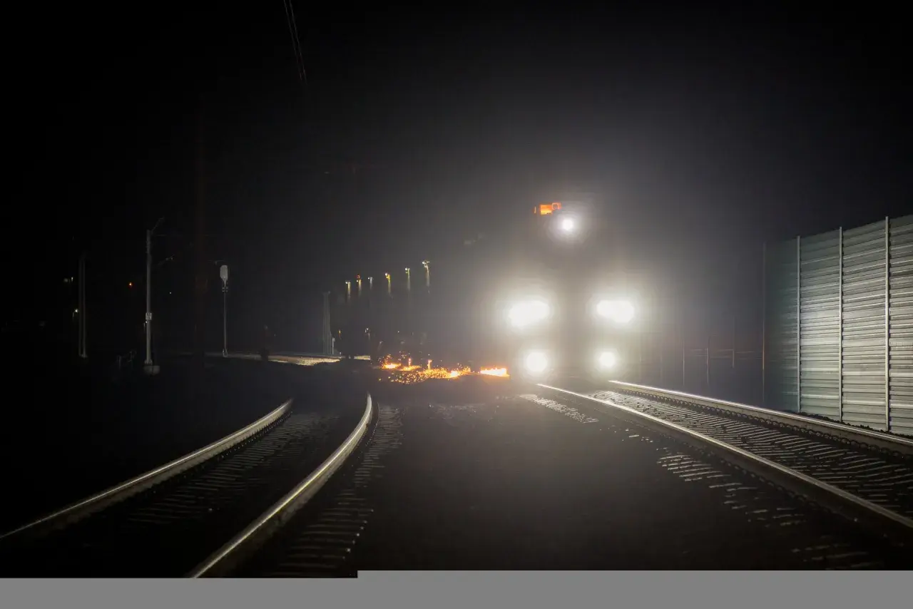 Nocny tor kolejowy rozświetlony reflektorami pociągu i iskrami z prac na torach.