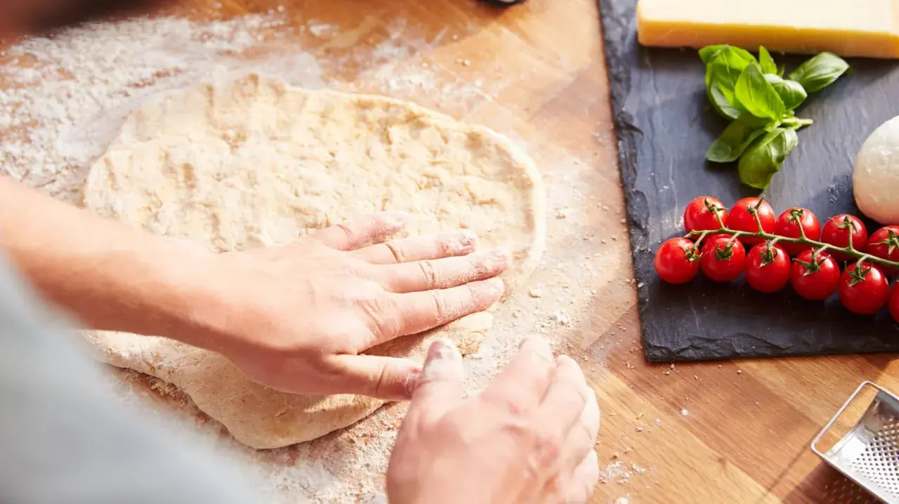 Hände formen den **pizzateig auftauen** auf einer bemehlten Holzoberfläche. Daneben liegen frische Tomaten, Mozzarella und Basilikum.
