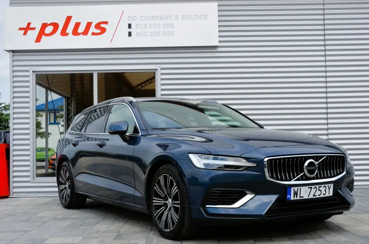 Ciemnoniebieskie Volvo V60 Hybrid z polskimi tablicami WL 7253Y przed budynkiem z logo "+plus".