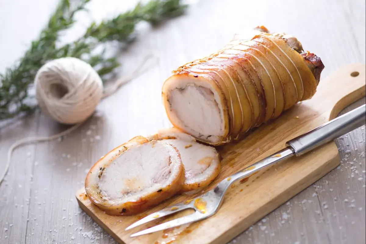 Ein aufgeschnittener, goldbraun gebratener Schweinebraten (Porchetta) auf einem Holzbrett mit Gabel und Rosmarin.