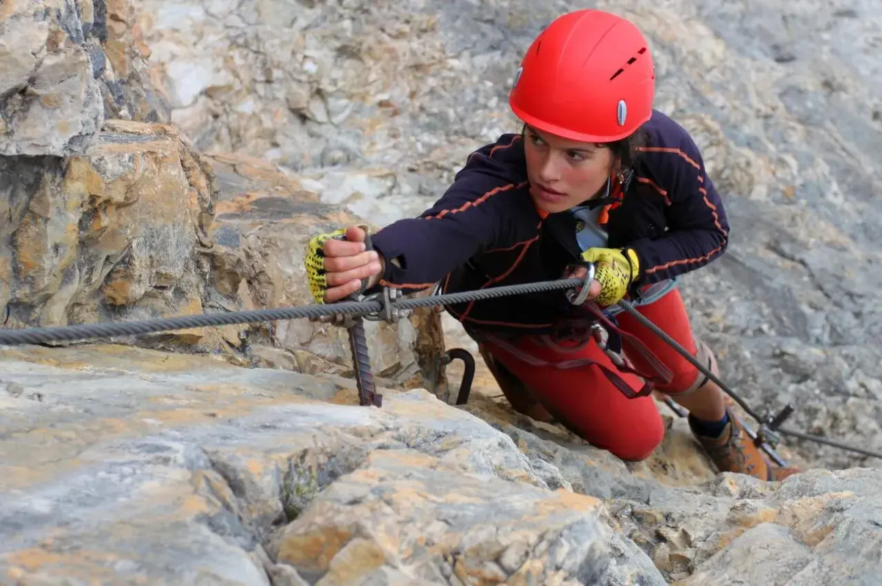 Młoda kobieta w kasku wspina się po skale, używając stalowej liny. To przygoda na via ferrata Czechy.