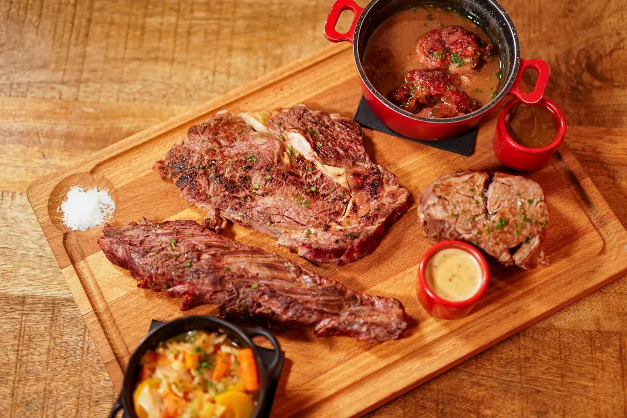 Un plateau en bois présente une entrecôte barbecue juteuse, des morceaux de viande braisée, des sauces et des légumes.