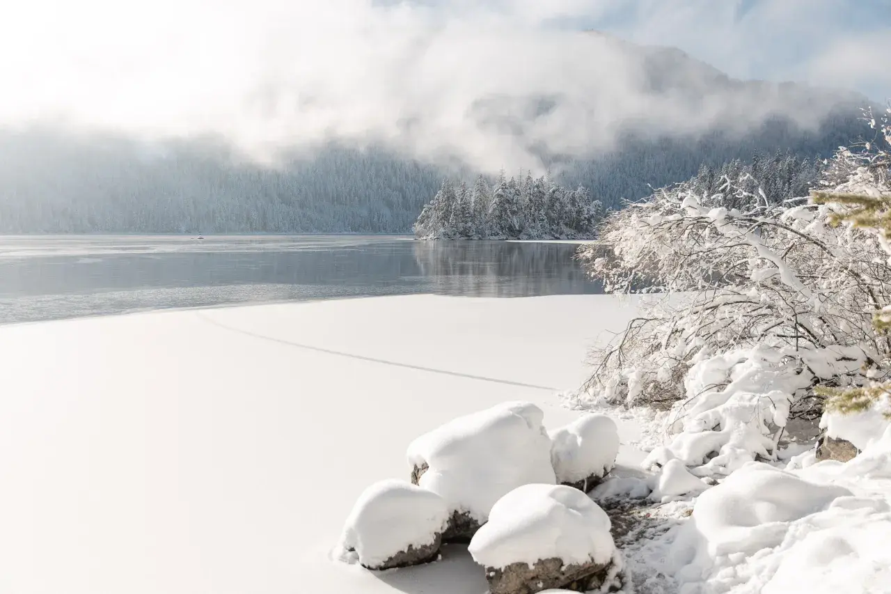 Eibsee Winter: Eisbedeckter See, schneebedeckte Bäume und Felsen unter nebligen Bergen. Ein friedlicher Anblick.
