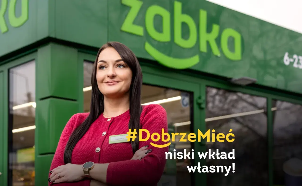 Uśmiechnięta franczyzobiorczyni Żabki przed sklepem. Hasło: #DobrzeMieć niski wkład własny!