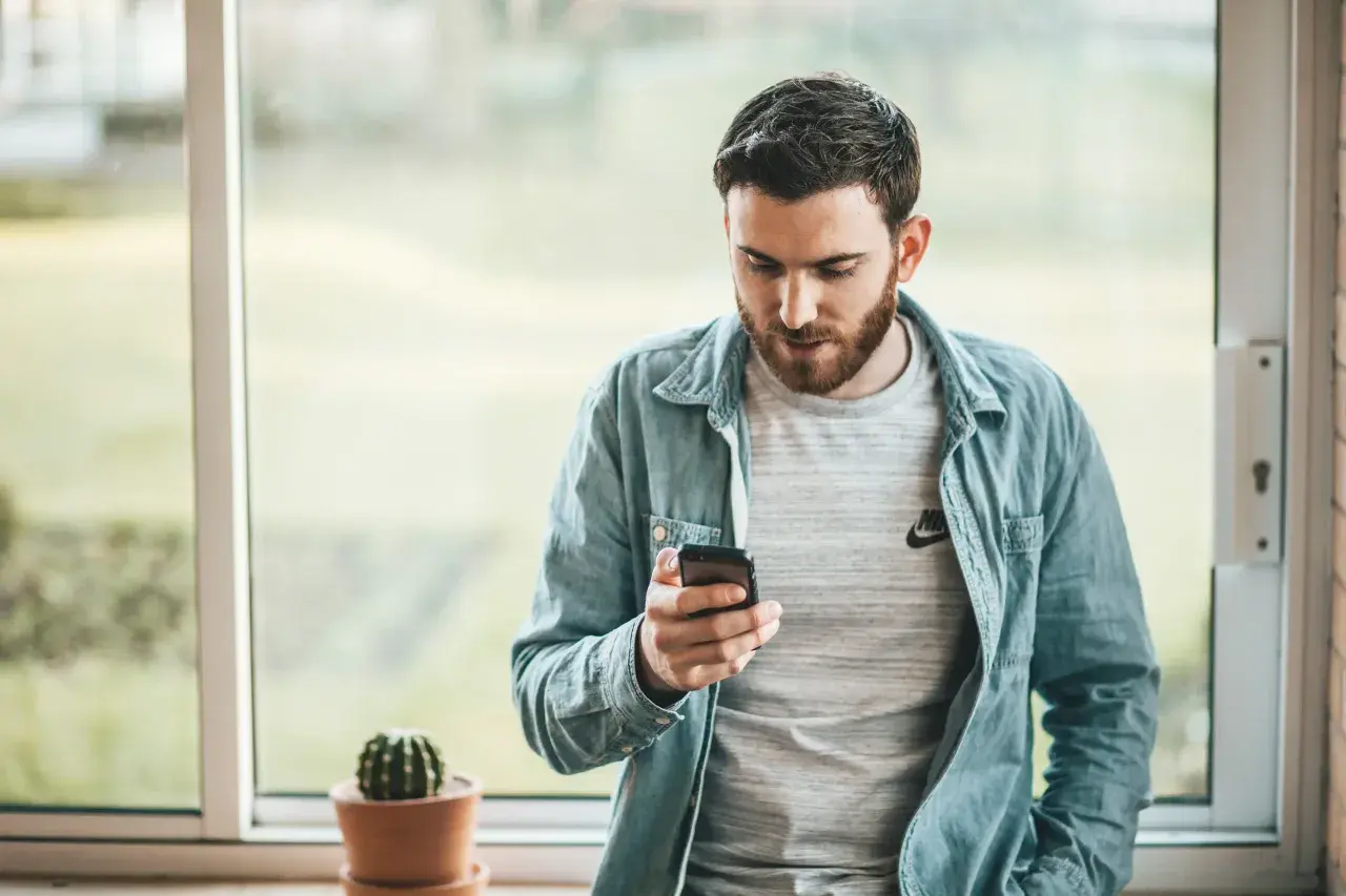 Hombre con barba mira su móvil, quizás buscando frases cuando no te responden. Un cactus en maceta decora el alféizar.