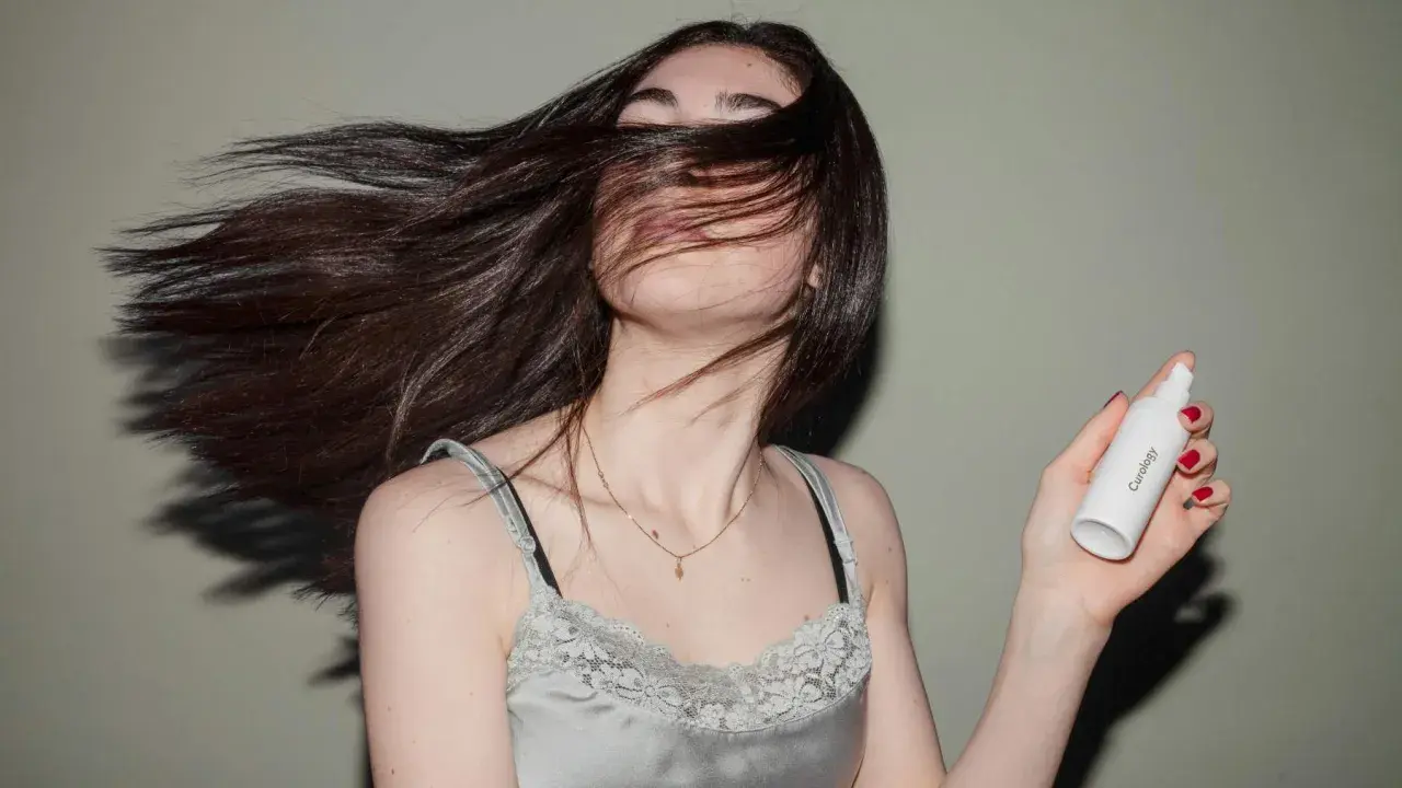 Mujer con cabello largo y oscuro moviéndose, sosteniendo un frasco de **aceite para el pelo**.
