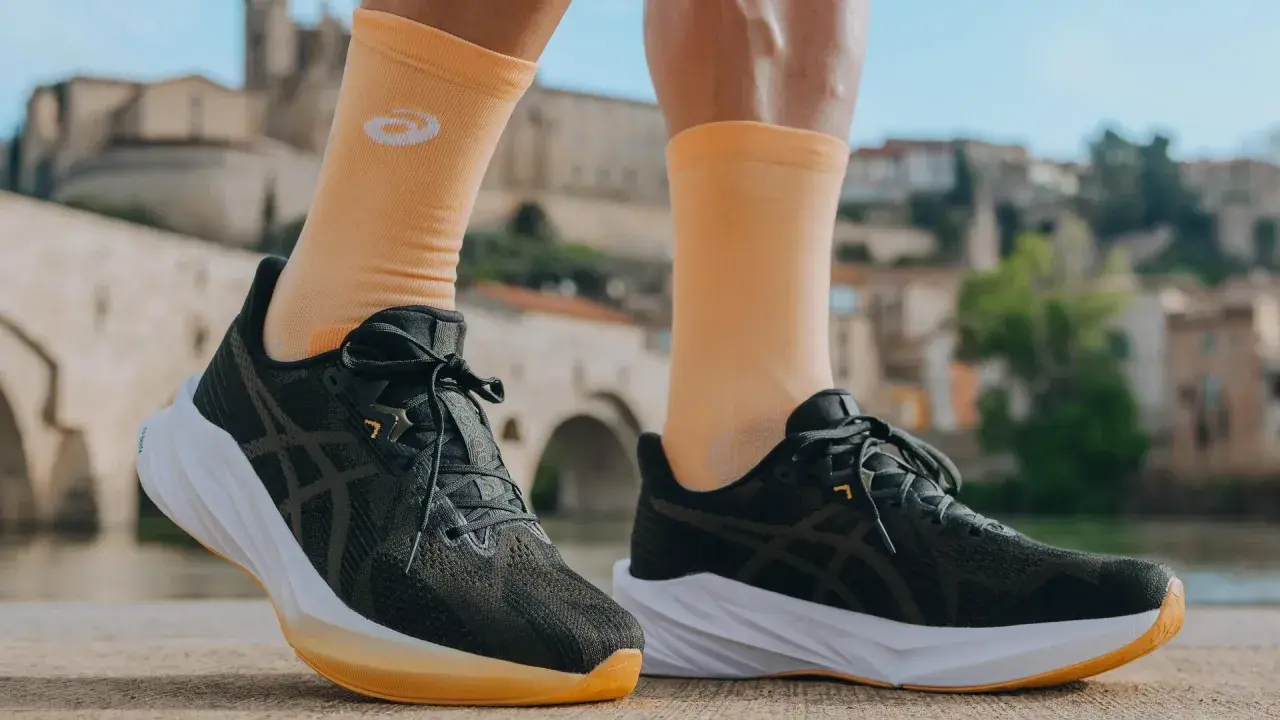 Biegacz w pomarańczowych skarpetkach z logo firmy Asics i ciemnych butach sportowych na tle mostu i zabudowań.