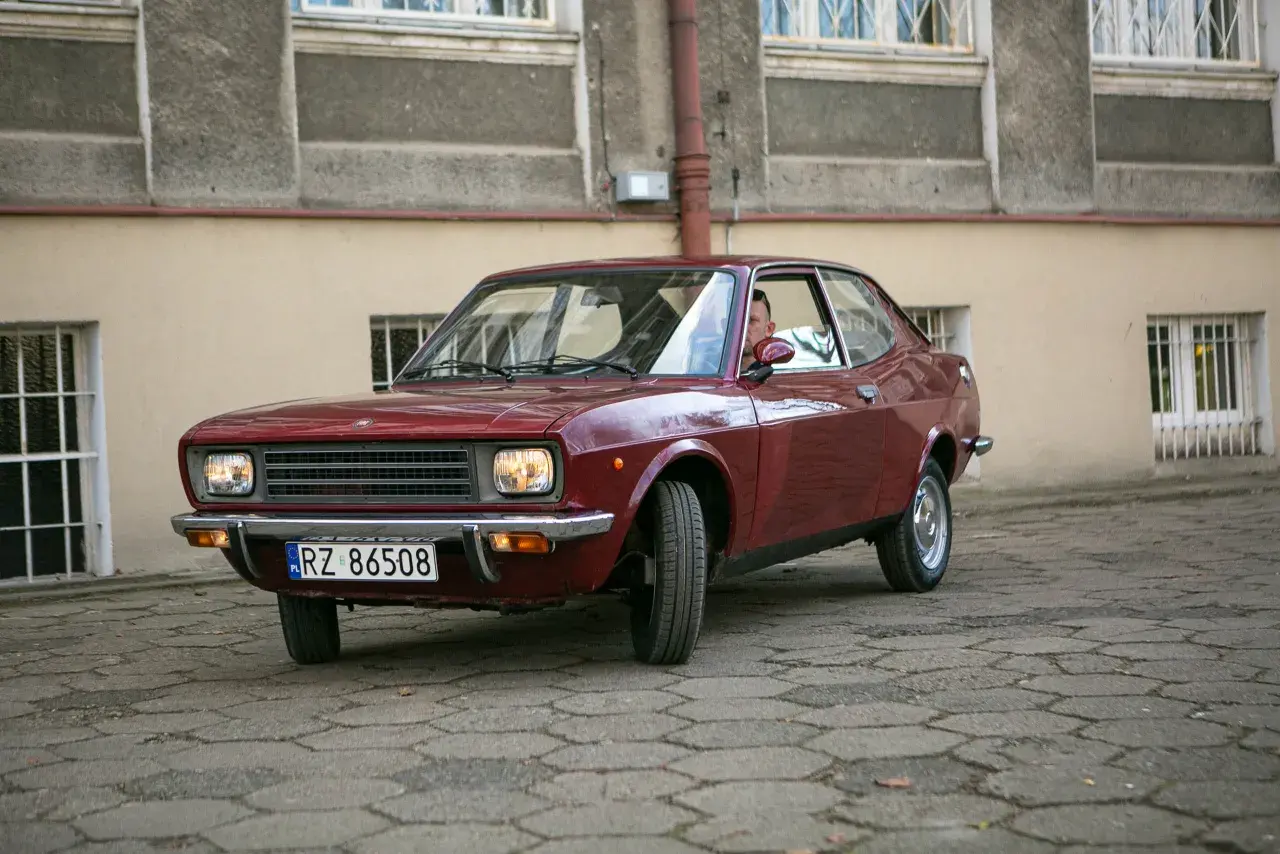 Czerwony fiat 128, klasyk z lat 70., zaparkowany na brukowanym dziedzińcu przed budynkiem.