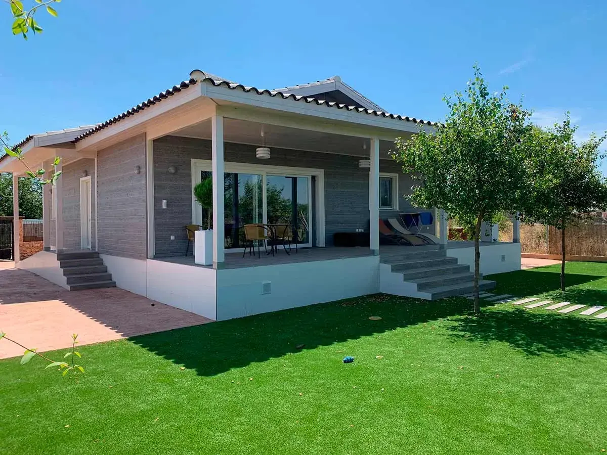 Moderna casa Canexel con porche, césped artificial y árboles frondosos bajo un cielo azul.