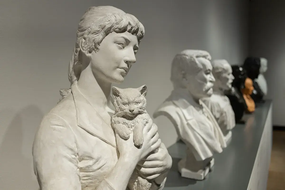 Rzeźba Camille Claudel "Plotkarki" - kobieta z kotem, obok popiersia mężczyzn.