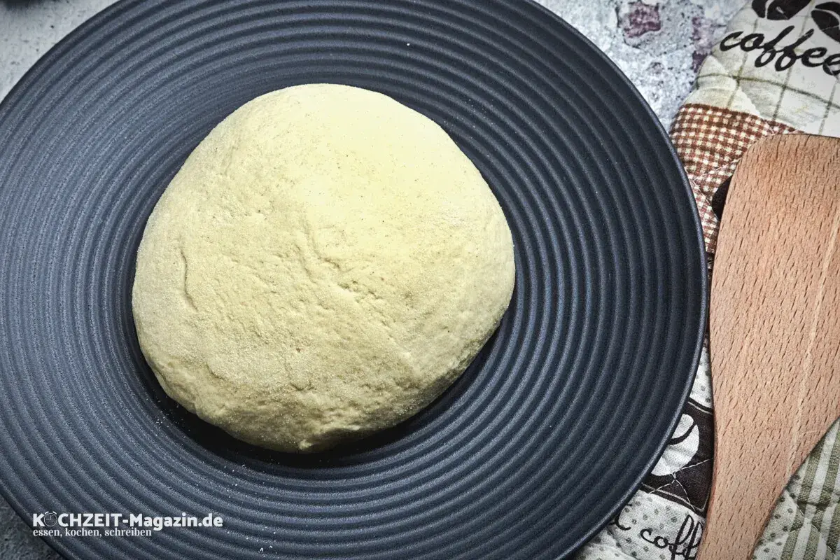 Frischer pizzateig auf schwarzer Platte, bereit für den pizzaofen. Ein Holzschaber liegt daneben.