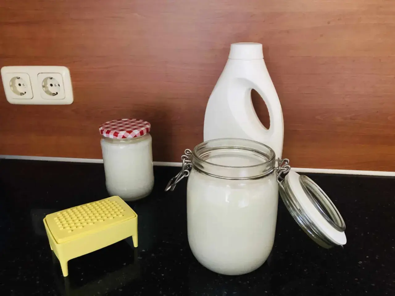 Zelf wasmiddel maken met dille en kamille. Glazen potten met wit wasmiddel, een gele rasp en een witte fles staan op het aanrecht.