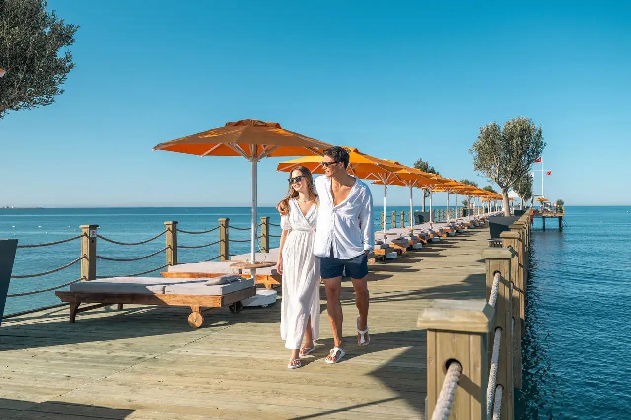 Ein Paar spaziert über einen Holzsteg am Meer, gesäumt von Liegen und Sonnenschirmen. Ein perfekter Urlaub im Arum Barut Collection.