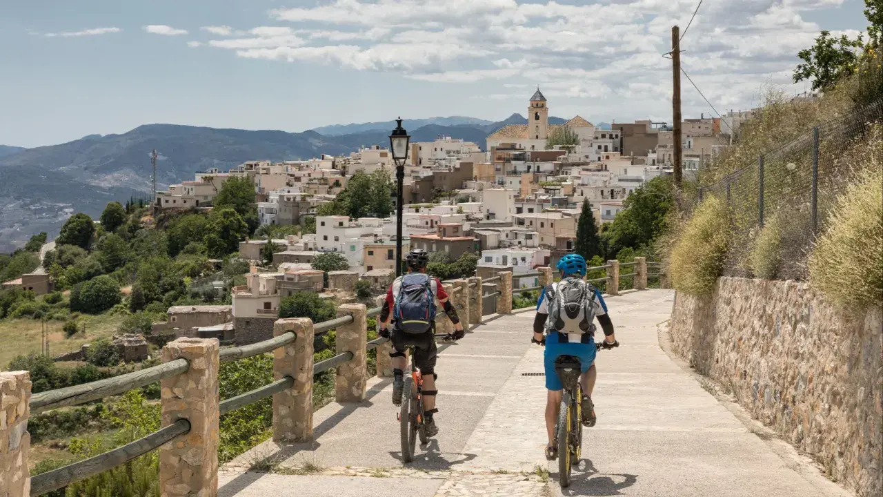 Dos ciclistas disfrutan de una ruta cicloturista ascendiendo hacia un pueblo blanco en la ladera de una montaña.