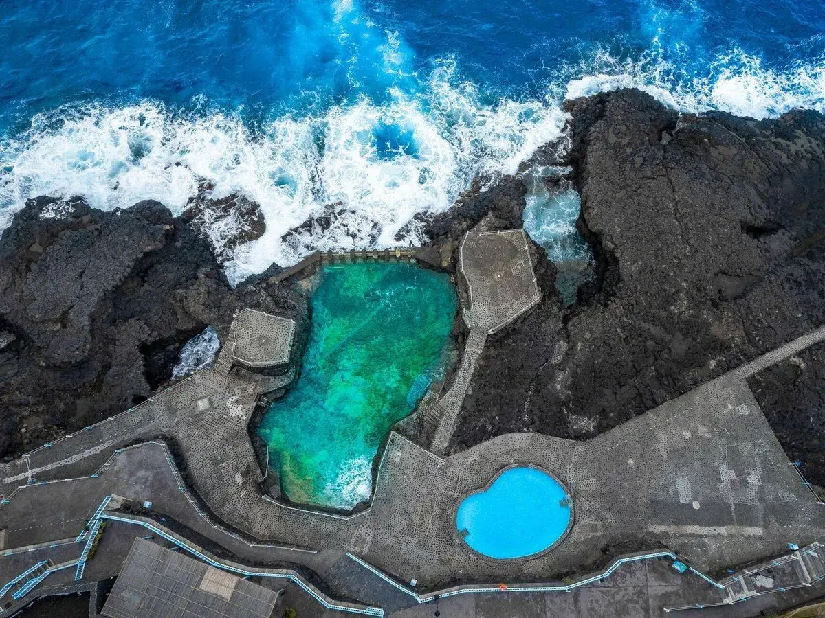 Piscinas naturales de agua turquesa y azul brillante, rodeadas de rocas volcánicas y el mar embravecido.