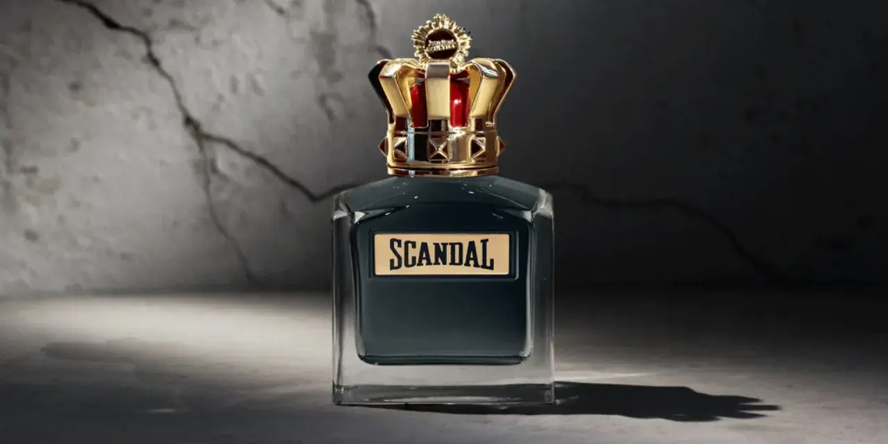 Elegancka butelka perfum Scandal męskie z koroną na nakrętce, stojąca na tle pękniętej ściany.
