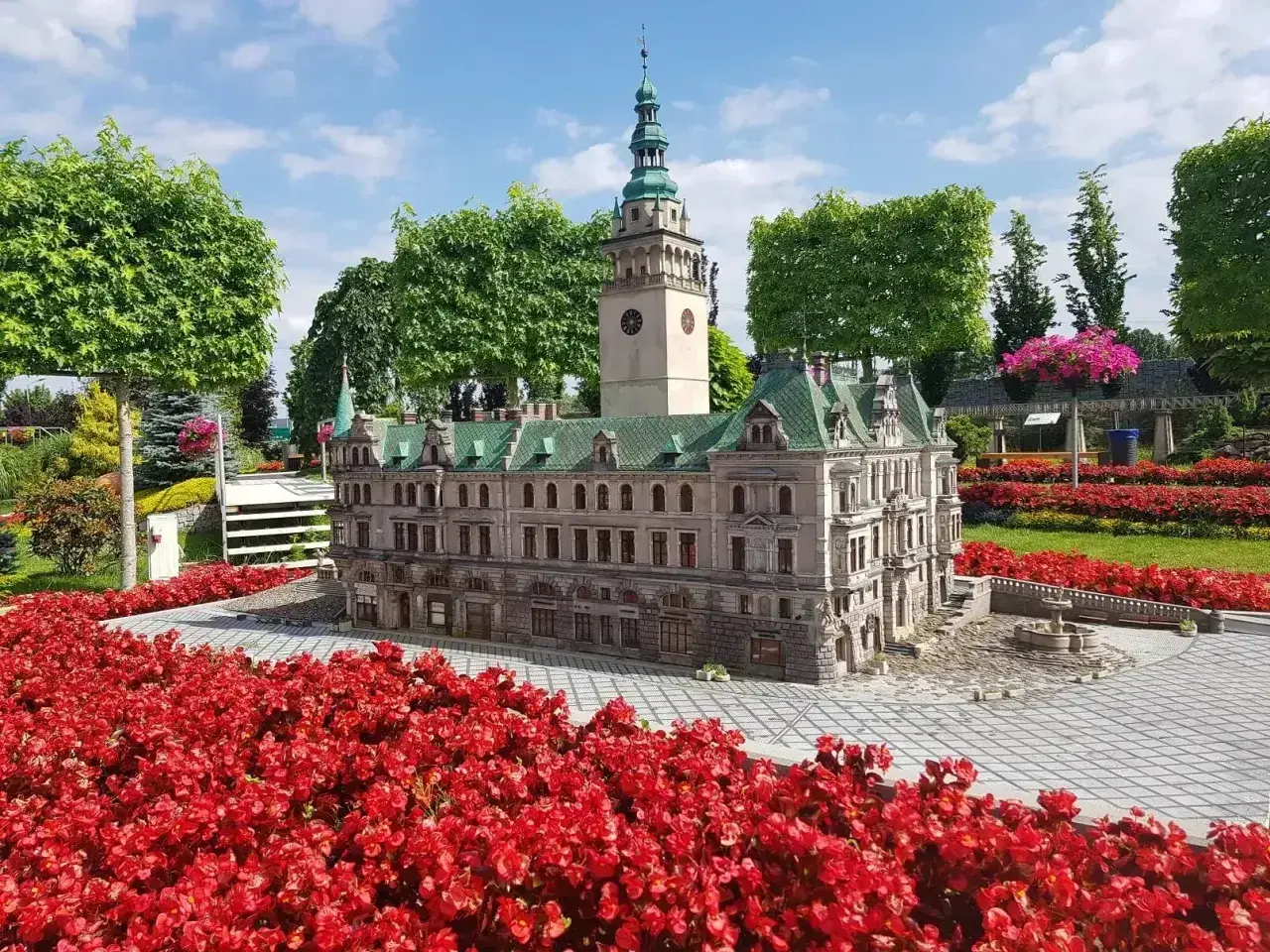 Miniatura ratusza z wieżą zegarową, otoczona czerwonymi kwiatami. Idealne miejsce na trasie po Kotlinie Kłodzkiej.