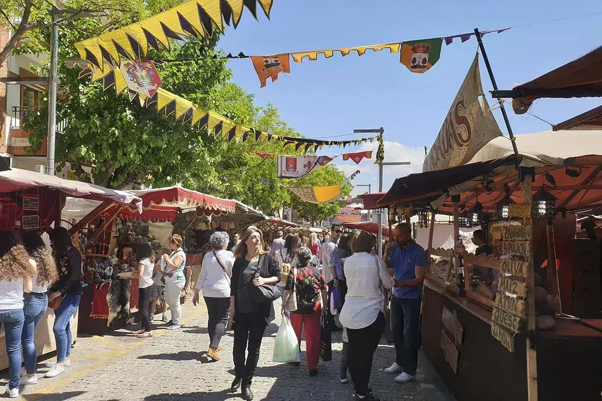 Gente pasea por el mercadillo medieval El Álamo, con puestos de artesanía y comida bajo banderines coloridos.