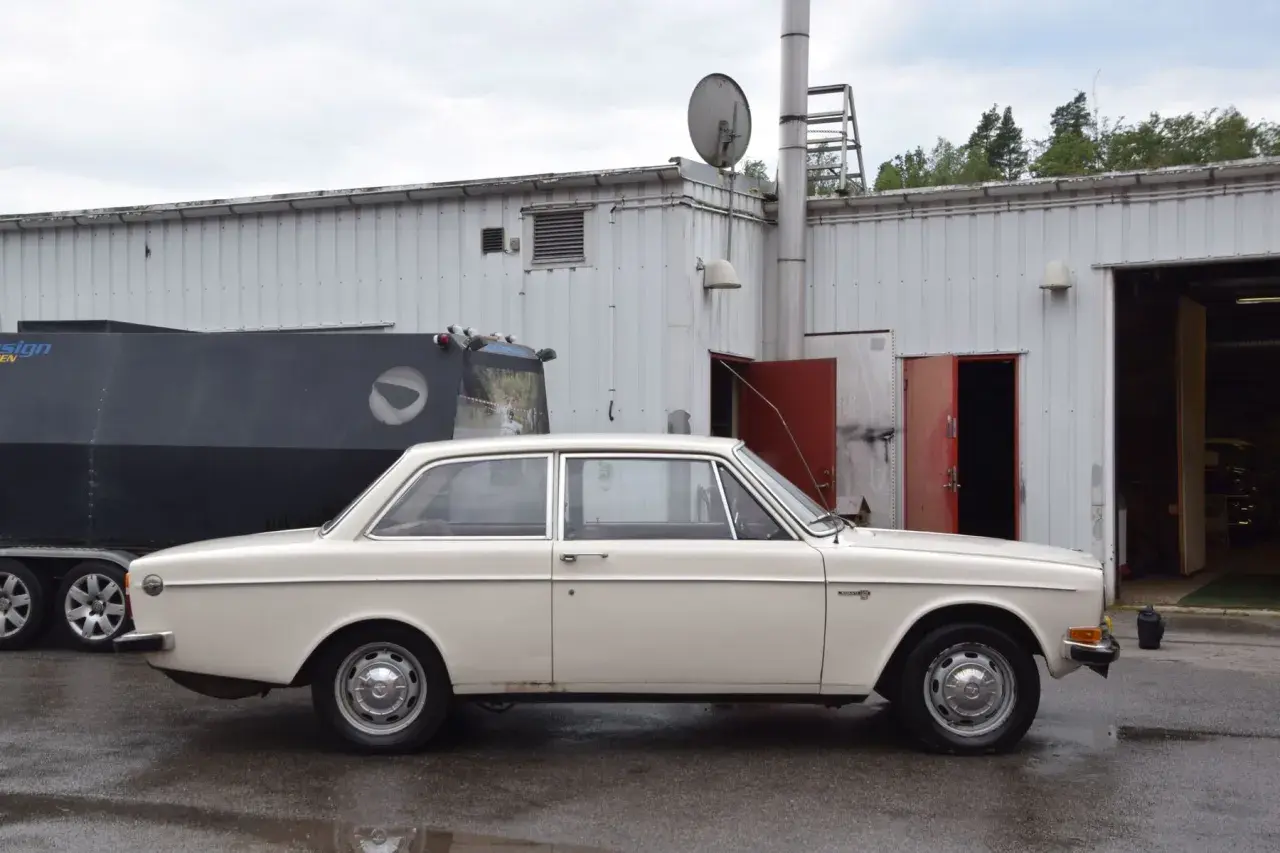 En vit Volvo 142 från 1973 står parkerad framför en industribyggnad.