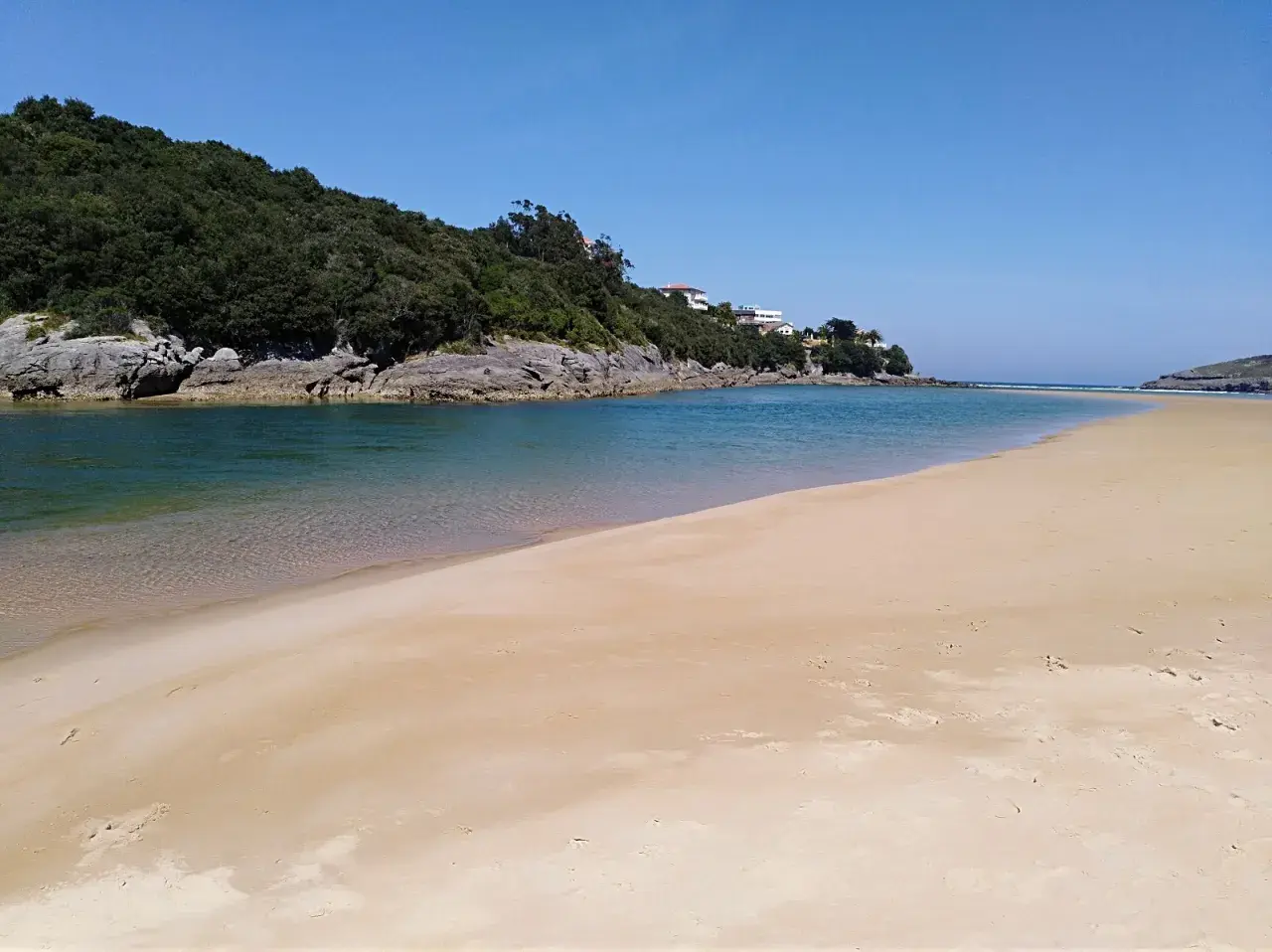 Amplia playa de la arena con aguas tranquilas y vegetación en la ladera. Un lugar ideal para los nudistas que buscan paz.