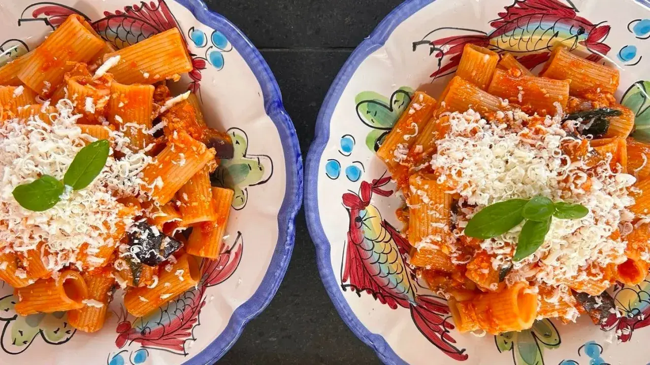 Deux assiettes de rigatoni alla norma, garnies de basilic frais et de fromage râpé, sur des plats décorés de poissons colorés.