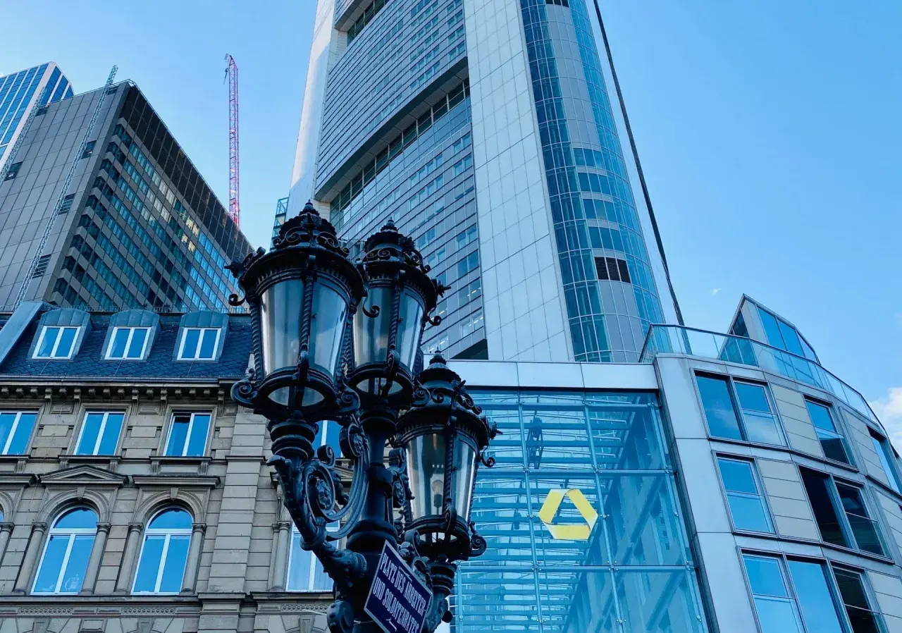 Ein historischer Laternenpfahl vor dem modernen Commerzbank-Gebäude in Frankfurt. Die Fassade spiegelt den blauen Himmel wider.