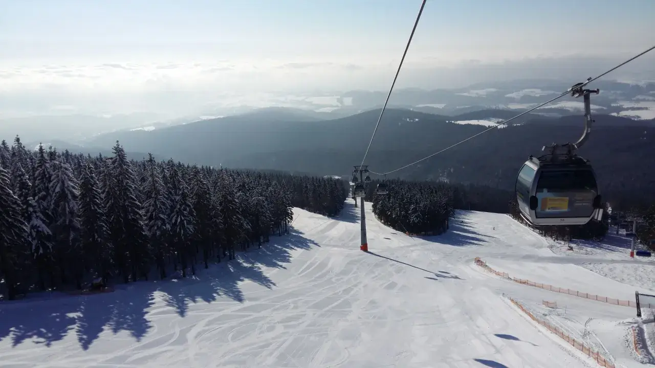 Trasa narciarska w Janskie Łaźnie, kolejka linowa wznosi się nad ośnieżonymi drzewami i stokiem.