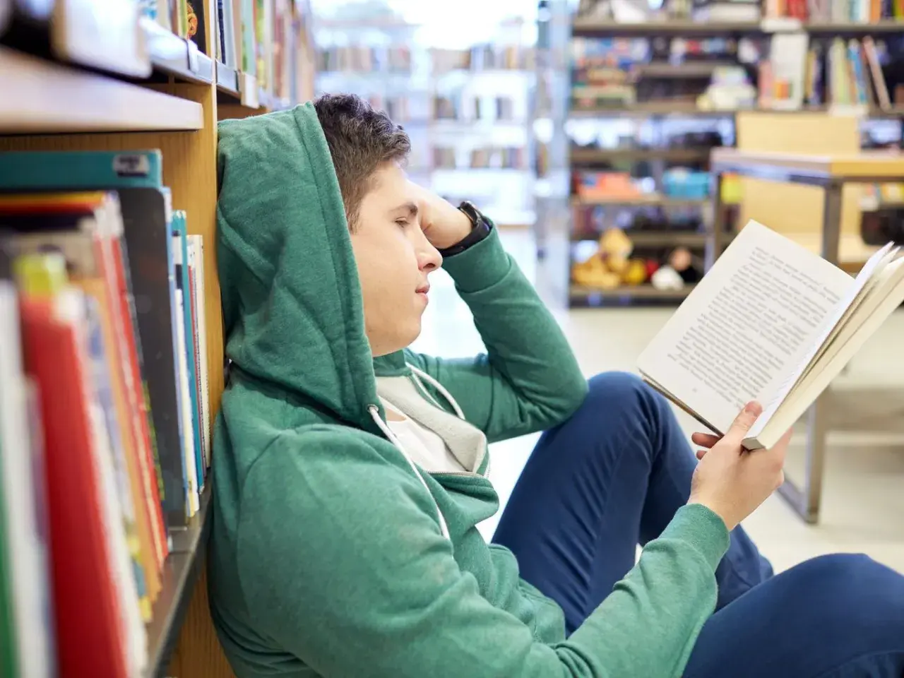 Chłopak w kapturze czyta książkę, szukając streszczenia makbeta. Siedzi na podłodze w bibliotece, opierając się o regał.