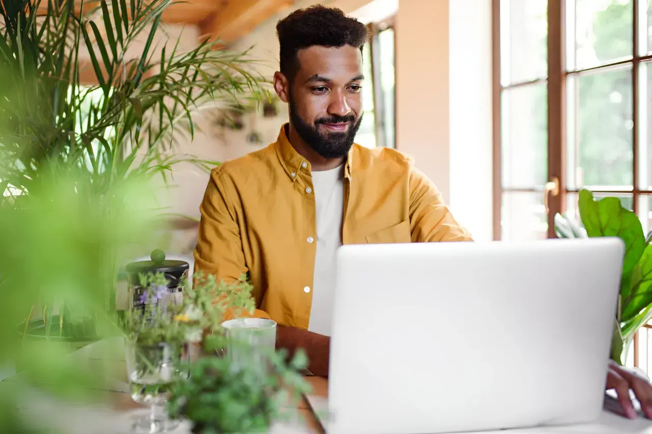 Uomo sorridente al laptop, circondato da piante. Sembra che stia imparando nuovi modi di dire per arricchire la sua comunicazione.
