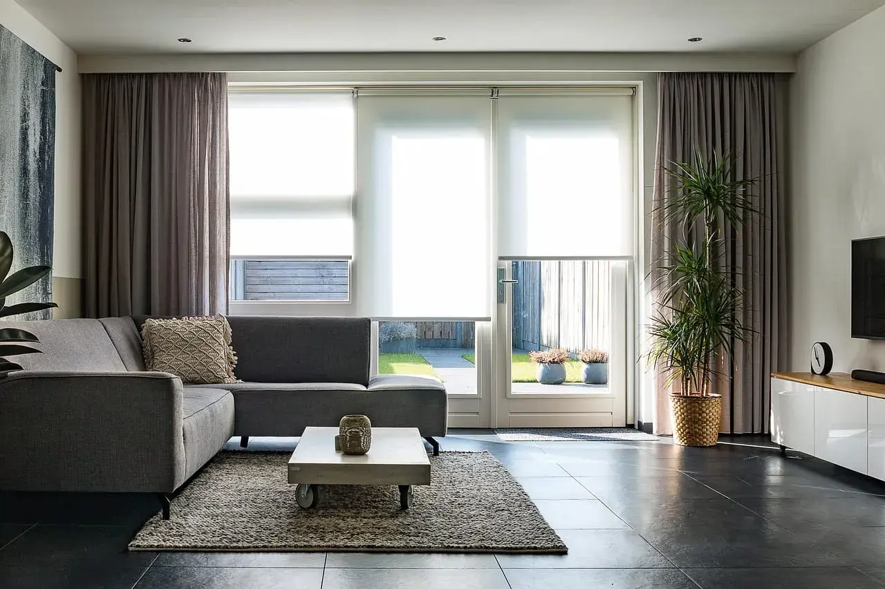Grå soffa i vardagsrummet med draperade gardiner och stora fönster som släpper in ljus.