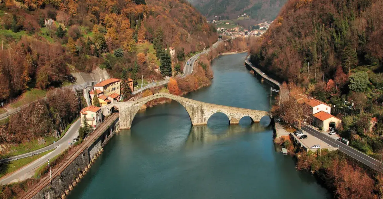 Il Ponte della Maddalena, con i suoi archi irregolari, attraversa un fiume circondato da colline autunnali e case colorate.