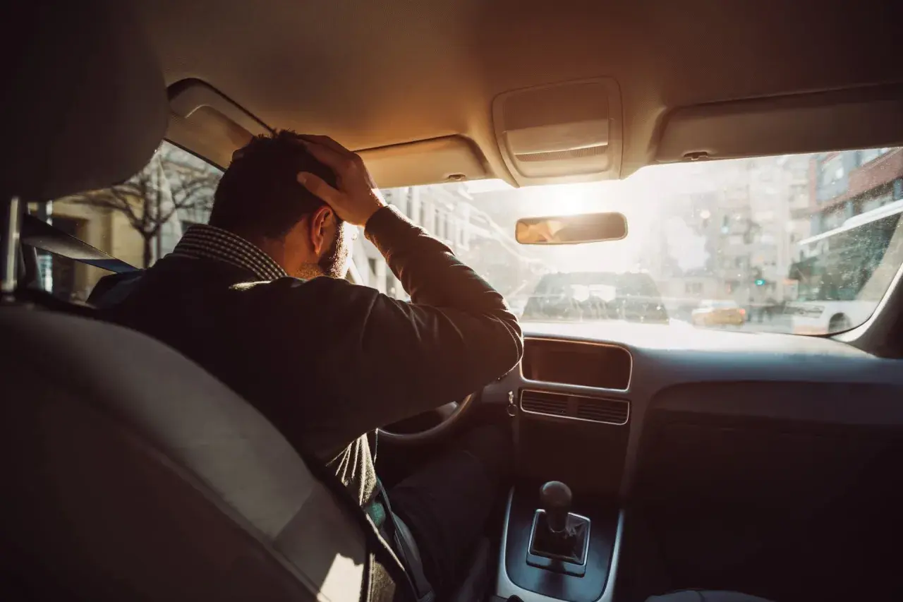 Hombre con mano en la cabeza dentro de un coche, buscando trucos para conducir sin nervios. El sol brilla a través del parabrisas.