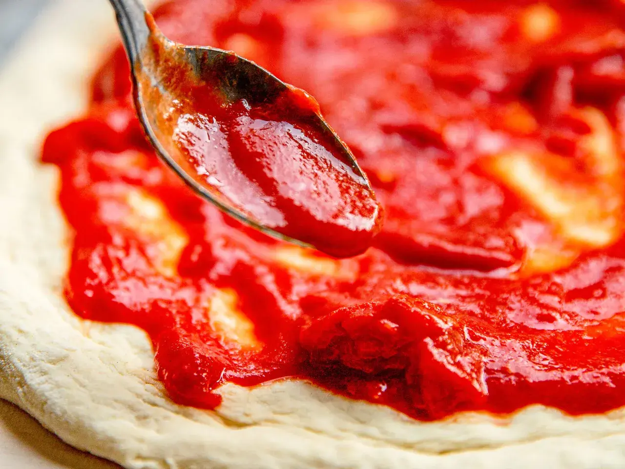 Eine Löffelspitze verteilt die leuchtend rote Pizza Sauce wie in der Pizzeria auf den Teig.