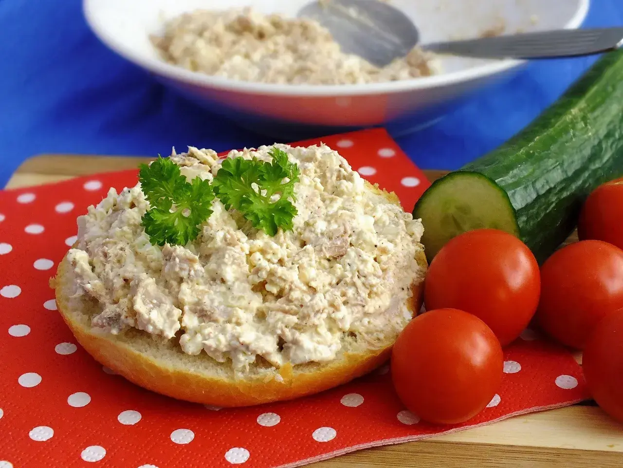 Pyszna pasta z tuńczyka i twarogu na bułce, ozdobiona natką pietruszki, obok pomidorki i ogórek.