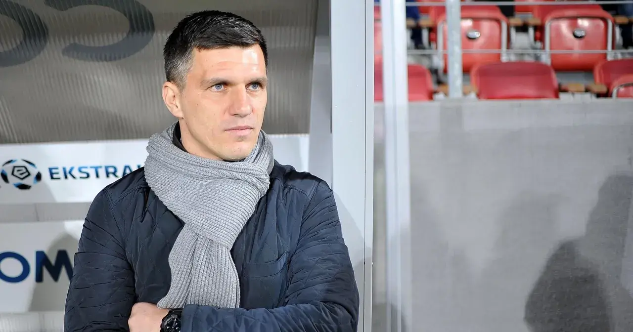 Robert Podoliński, trener w szaliku, spogląda w bok na tle stadionu z czerwonymi siedzeniami.