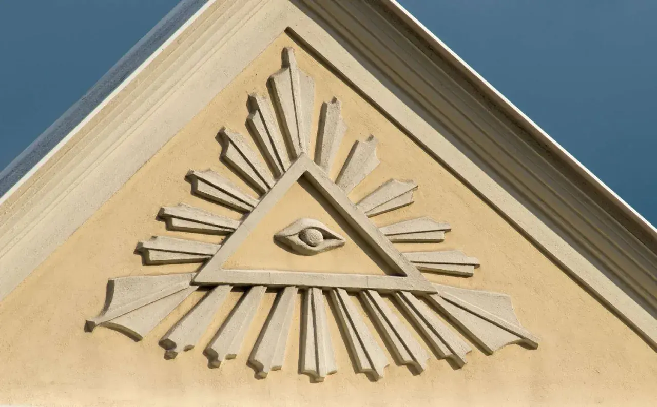 Kamienna płaskorzeźba oka w trójkącie z promieniami na żółtej ścianie budynku.